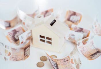 Buying a house in cash UK. Wooden House model, piggy bank with British currency on the white table.